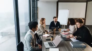 Gruppo in riunione attorno a un tavolo con laptop e appunti.