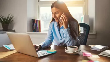 Persona al telefono mentre lavora al computer portatile in un ufficio illuminato dal sole.
