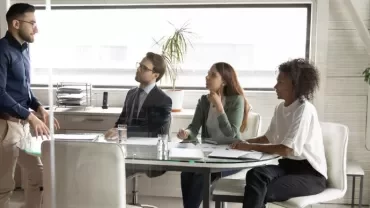 Persona in piedi di fronte a tre persone sedute in una sala riunioni.