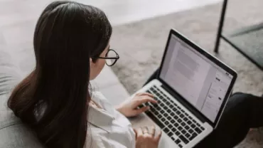 Persona con occhiali lavora al computer portatile su un divano.