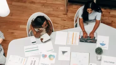 Persone al lavoro in un ufficio con documenti e laptop su un tavolo.