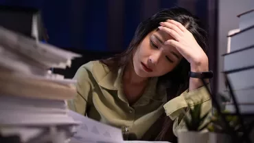 Persona al tavolo sommersa da documenti, sembra concentrata e pensierosa.