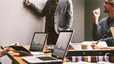 Riunione in ufficio con persone che discutono e laptop aperti sul tavolo.