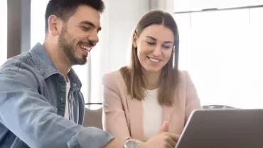 Due persone sedute ad un tavolo sorridono davanti al PC