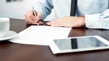 Persona che scrive su un documento con penna, tavolo con tazza e tablet.