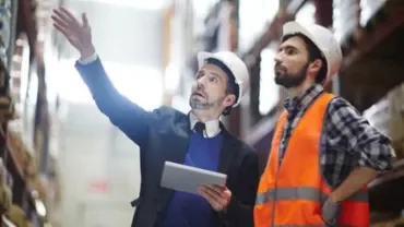 Due persone con caschi da cantiere in un magazzino, una indica, l'altra osserva.