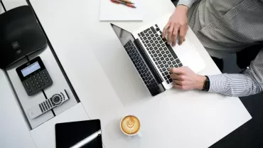 Persona lavorando al computer con caffè, telefono e tablet su un tavolo bianco.