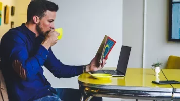 Persona seduta con tazza, libro aperto e laptop su tavolo giallo