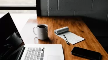 Scrivania con laptop, tazza di caffè, taccuino e penna.