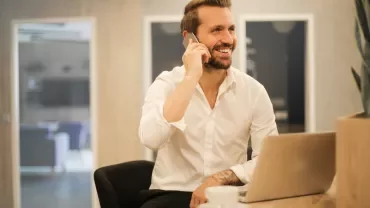 Persona sorridente al telefono, seduta a un tavolo con laptop e caffè.
