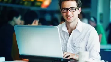 Persona sorridente seduta al tavolo con un laptop in un ambiente interno.