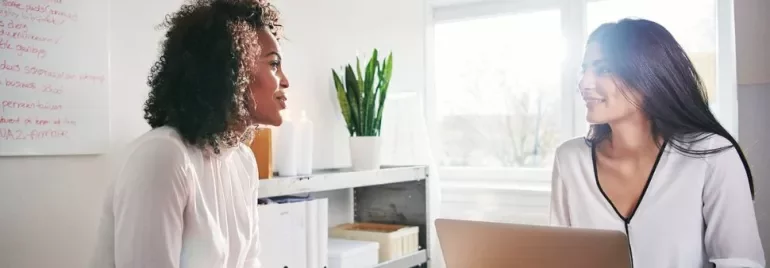 Due persone conversano in un ufficio luminoso con un laptop aperto sul tavolo.
