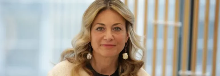 Persona sorridente con capelli ondulati e sfondo di finestre e legno.