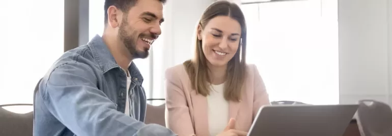 Due persone sedute ad un tavolo sorridono davanti al PC