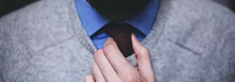 Persona sistema una cravatta rossa su camicia blu e maglione grigio.