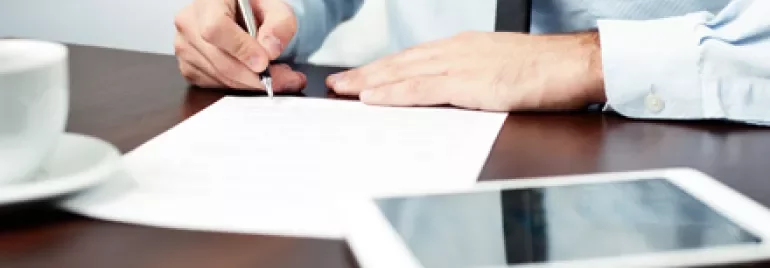 Persona che scrive su un documento con penna, tavolo con tazza e tablet.