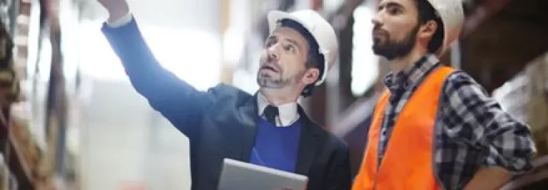 Due persone con caschi da cantiere in un magazzino, una indica, l'altra osserva.