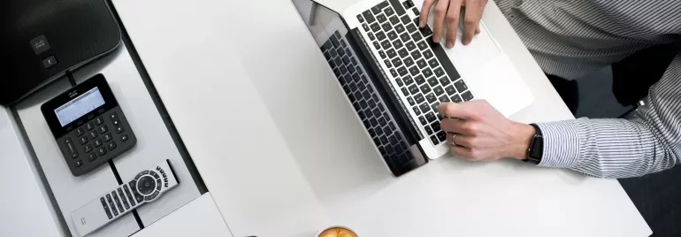 Persona lavorando al computer con caffè, telefono e tablet su un tavolo bianco.