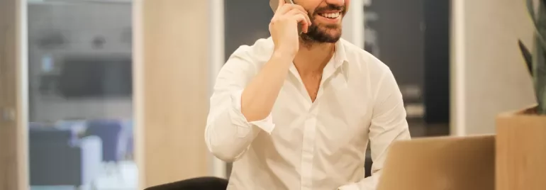 Persona sorridente al telefono, seduta a un tavolo con laptop e caffè.