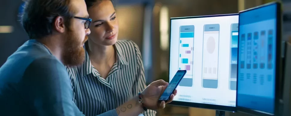 Due persone analizzano interfacce app su schermo e telefono.