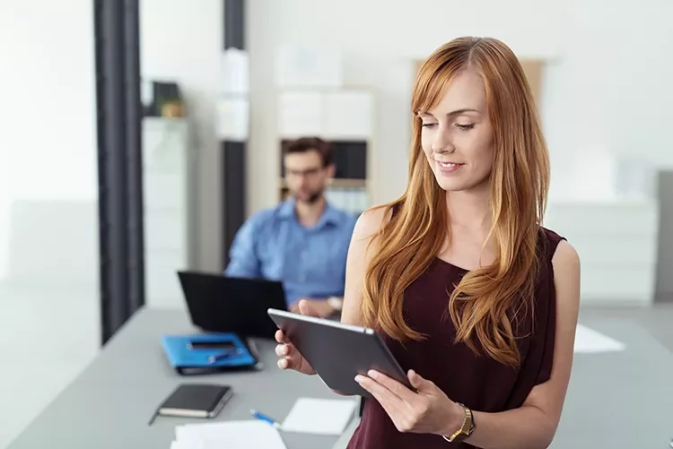 Persona in ufficio con tablet, sfondo sfocato con collega al computer.