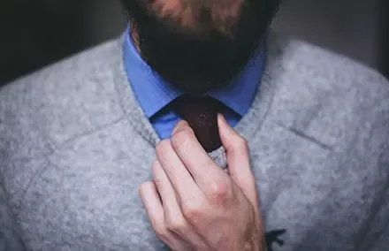 Persona sistema una cravatta rossa su camicia blu e maglione grigio.