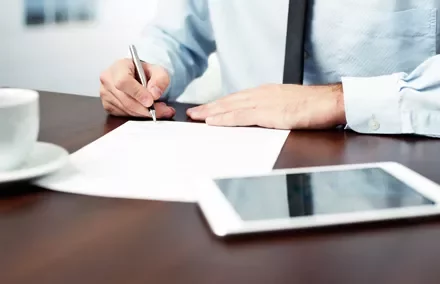 Persona che scrive su un documento con penna, tavolo con tazza e tablet.