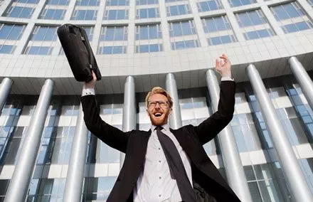 Persona sorridente solleva una valigetta davanti a un edificio moderno.