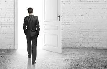 Persona in abito si allontana verso una porta aperta, luce intensa all'esterno.