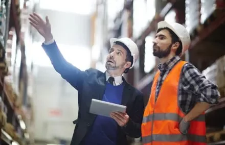 Due persone con caschi da cantiere in un magazzino, una indica, l'altra osserva.