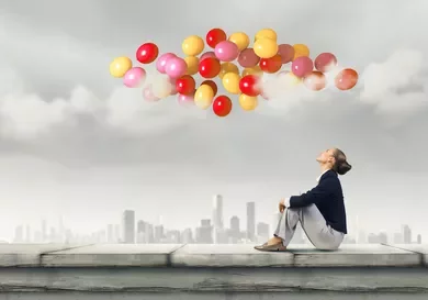 Persona seduta su un tetto osserva palloncini colorati nel cielo grigio.