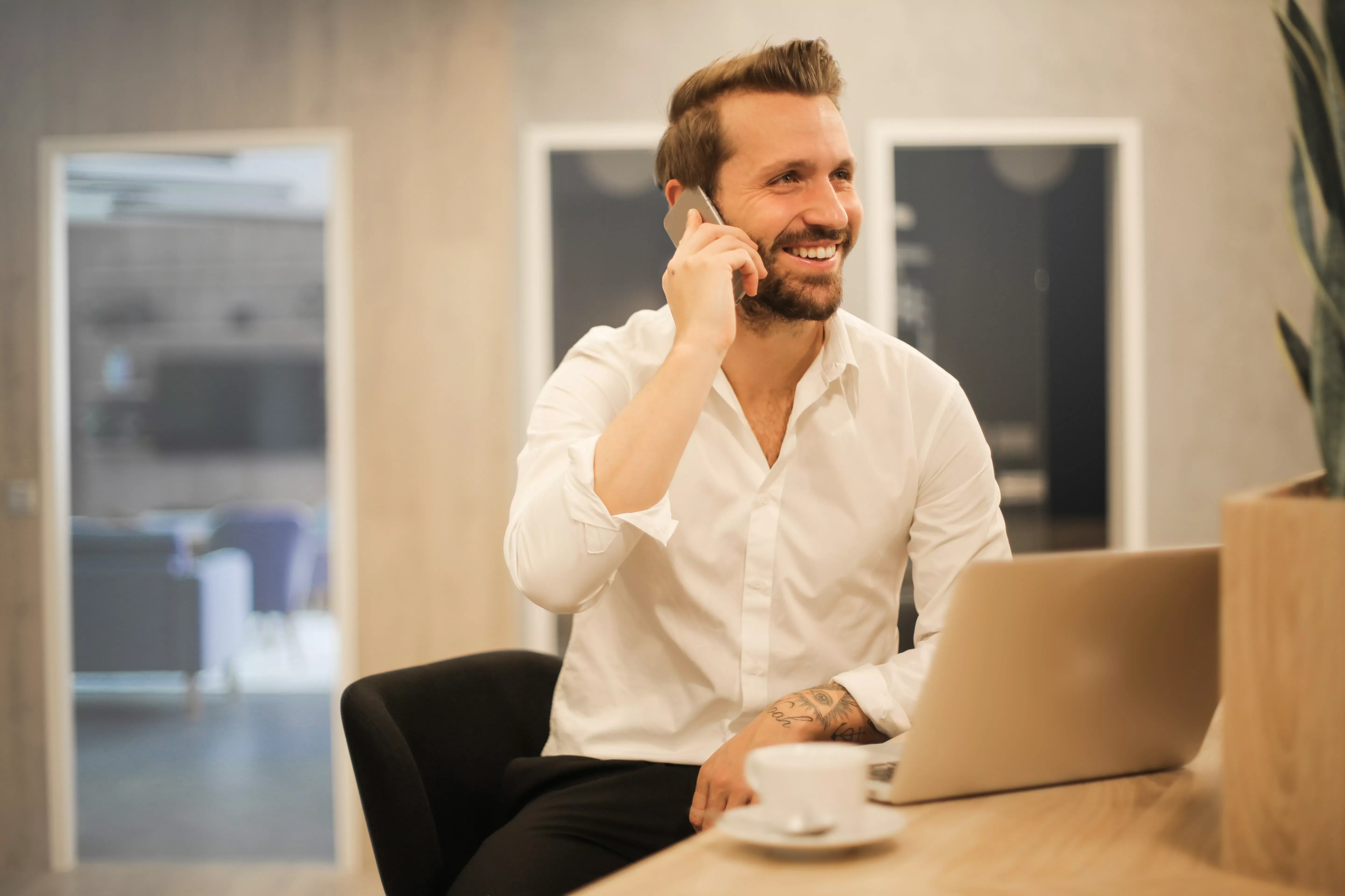 Persona sorridente al telefono, seduta a un tavolo con laptop e caffè.